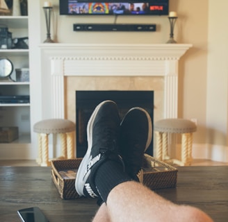 A person is relaxing in a living room with feet up on a wooden table. A TV mounted above a fireplace is displaying a popular streaming platform. Shelves on either side of the fireplace hold decorative items. A smartphone can be seen on the table.