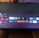 A television screen displays a smart TV interface with multiple app icons arranged in rows. Options such as Settings, Sources, Home, Search, and TV are listed on the left side. Some recognizable app icons include Netflix, Prime Video, BBC iPlayer, ITV Hub, All 4, My5, YouTube, and TikTok. The time and date are shown in the top right corner.