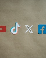 Four social media platform logos are arranged on a brown background. From left to right, the logos are for YouTube in red, TikTok in a combination of red, blue, and white, X in white, and Facebook in blue.
